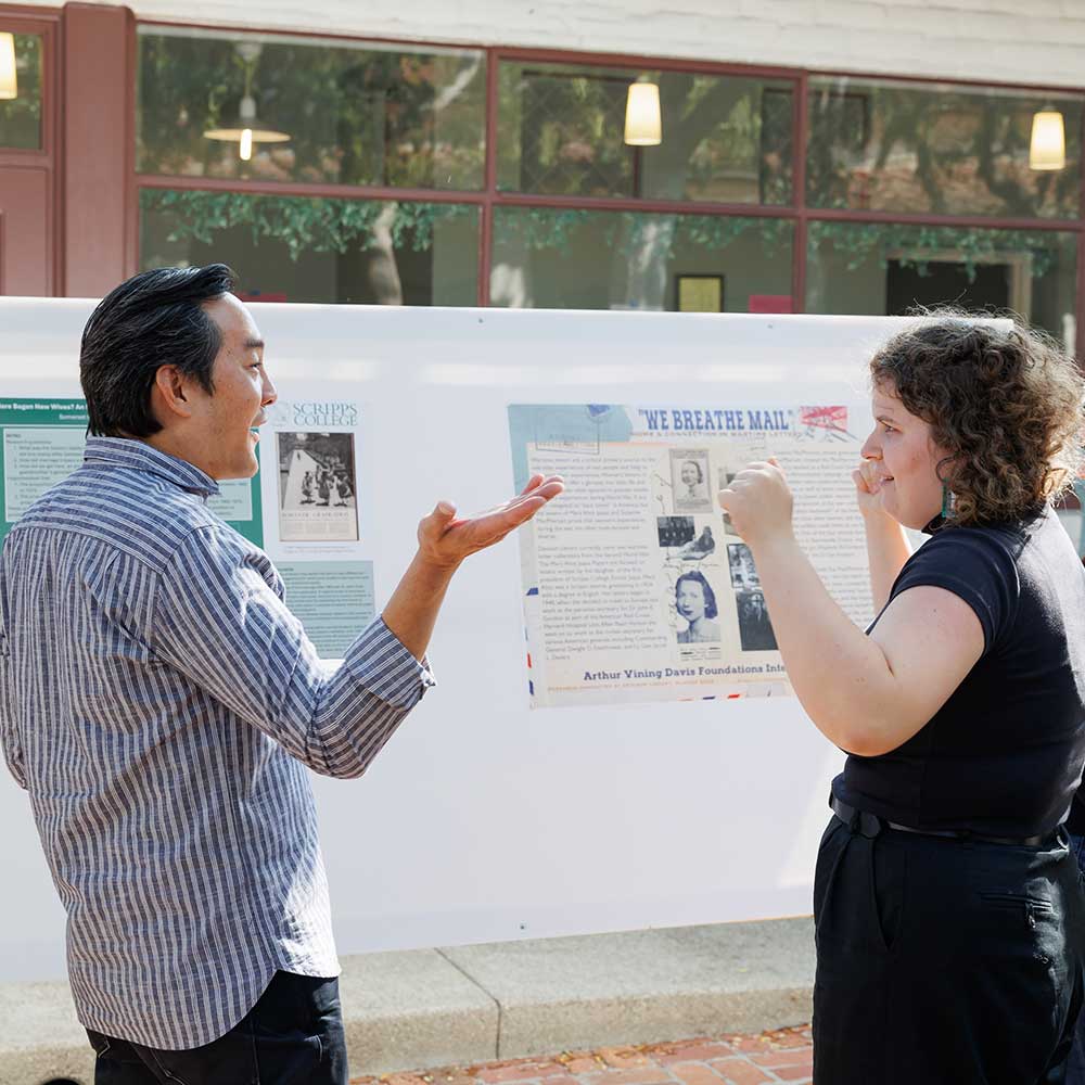 Professor Warren Liu, an Asian man, speaks with Skye Curren, a white female student, in front of a poster board.