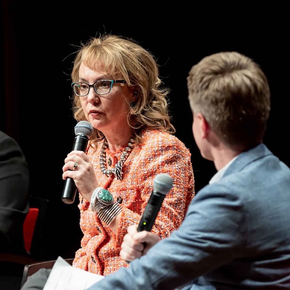 Gabby Giffords speaks into a microphone on stage at Scripps. A male interviewer in a blue suit sits next to her.