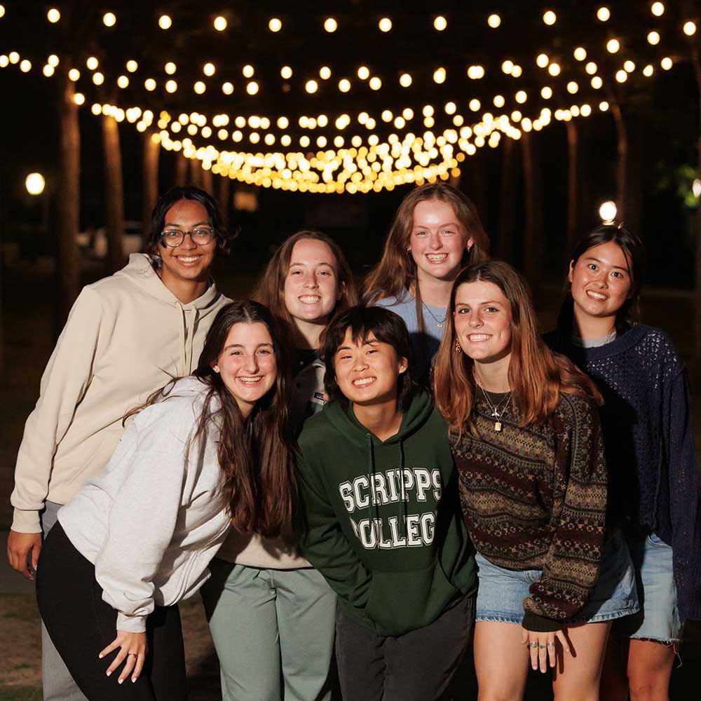 Snapshot of several Scripps College students smiling at the camera. String lights glow behind them.